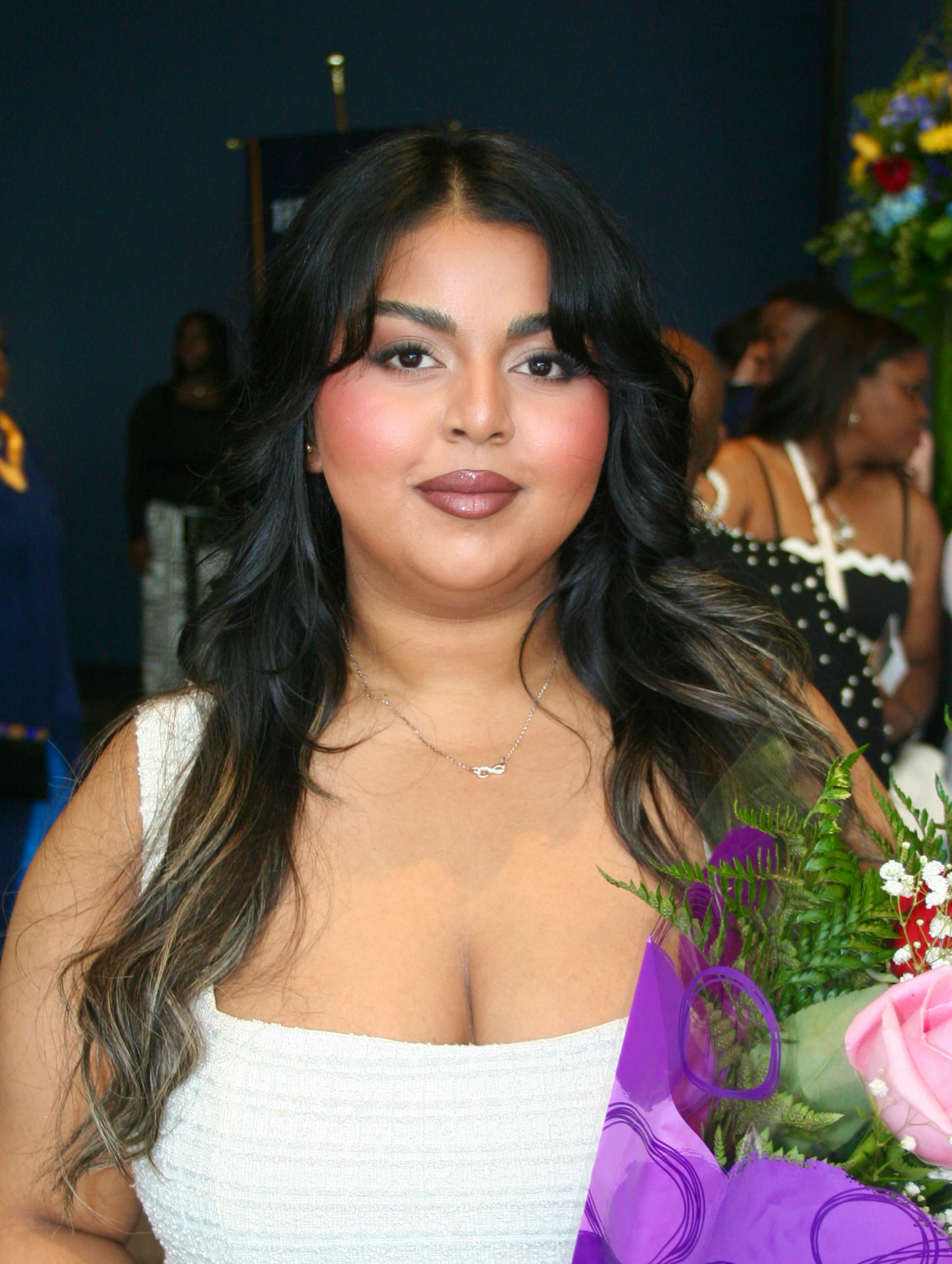 Rachel holding a bouquet of purple and pink flowers, wearing a white textured top and a mauve soft glam makeup look